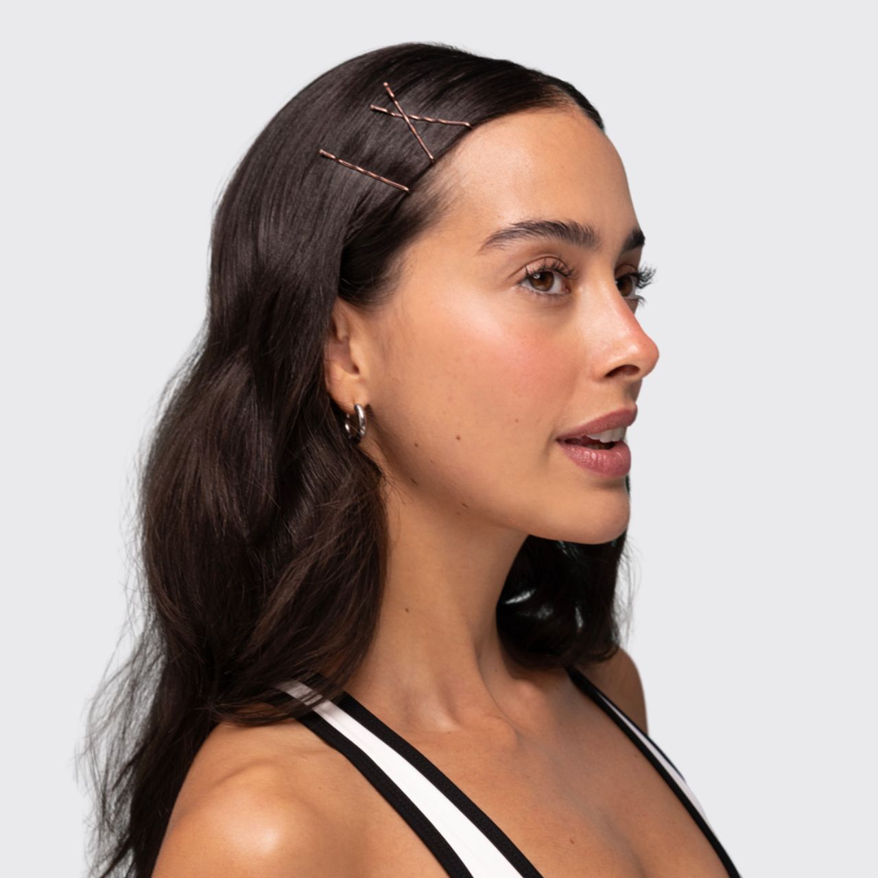 A woman with long dark brown hair wearing three Kitsch blonde bobby pins on the side of her hair, shown in a side profile against a light gray background.