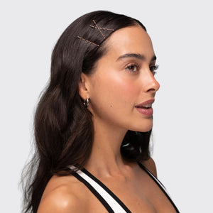 A woman with long dark brown hair wearing three Kitsch blonde bobby pins on the side of her hair, shown in a side profile against a light gray background.