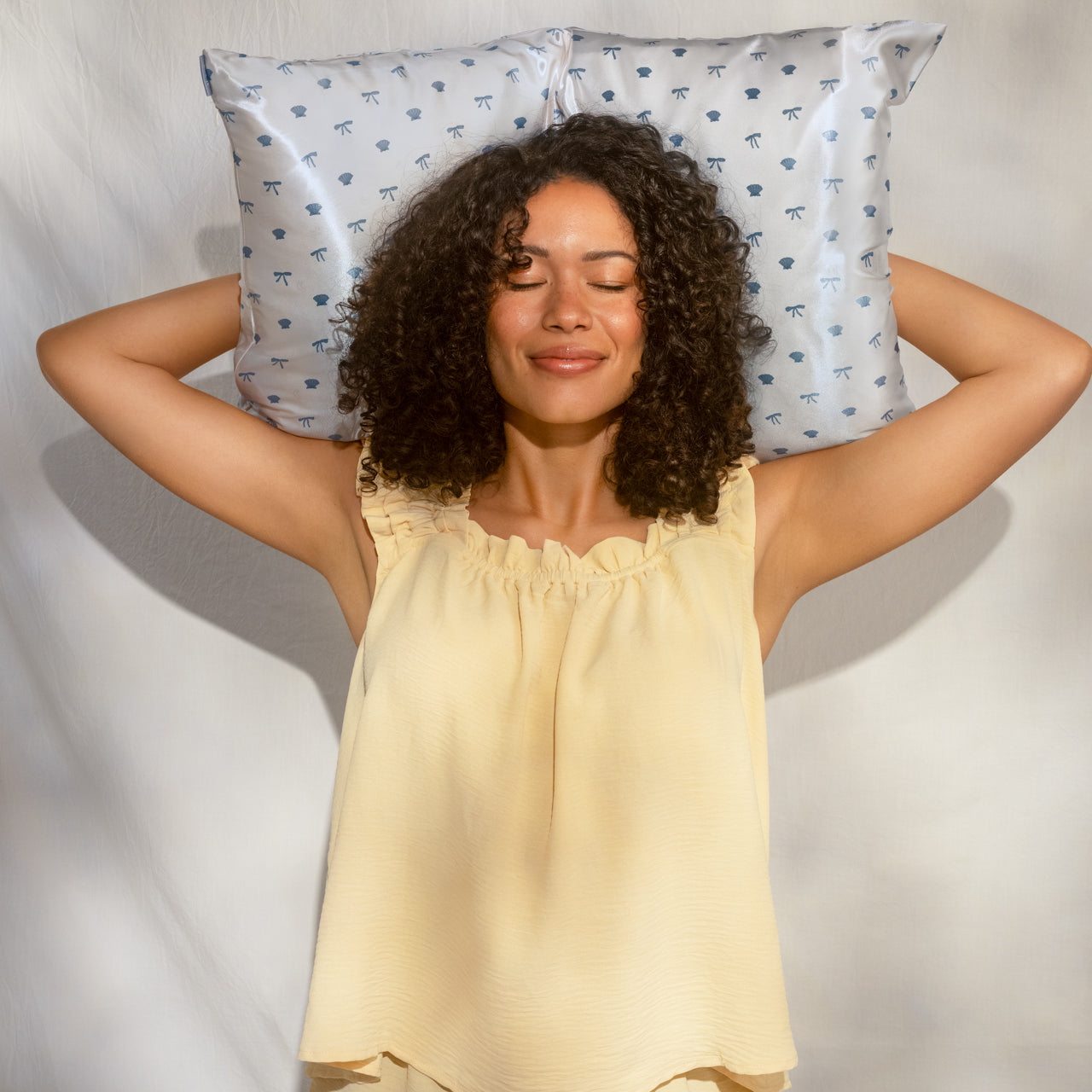 Female model with curly hair in yellow top lying on KITSCH Satin Pillowcase in Pearlized Cream & Coastal Blue Print — eyes closed, arms raised behind head, serene lifestyle portrait — Reduces friction against hair & skin, helps reduce breakouts & keeps skin feeling hydrated