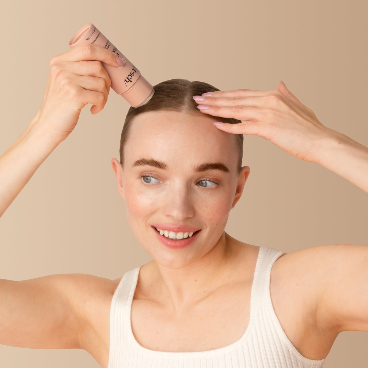 KITSCH Hair Wax Stick — lifestyle portrait of female model with light hair pulled back, applying stick at hairline with both hands, smiling, white tank top, warm beige background — Tames stubborn strands for a sleek & polished style — Effortlessly smooths hair with a convenient solid stick design for touch-ups on the go — All-day hold without texture or greasiness