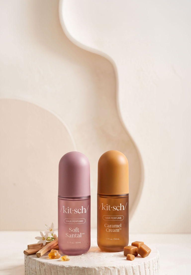 Two Kitsch hair perfume bottles, one in Soft Santal (dusty mauve) and one in Caramel Cream (caramel brown), displayed on a textured stone pedestal with decorative cinnamon sticks and caramel pieces against a warm beige background.
